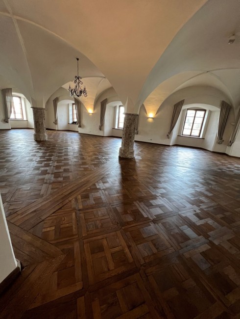 Großer Raum mit Holzboden, gewölbter Decke, Kronleuchter und Fenstern mit Vorhängen